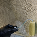 Black-gloved hand rolling resin into biaxial fiberglass laid across a hull side, the diagonal fiber pattern wetting out under a bent-handle roller