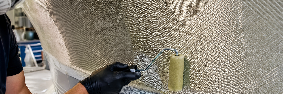 Black-gloved hand rolling resin into biaxial fiberglass laid across a hull side, the diagonal fiber pattern wetting out under a bent-handle roller