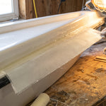 Gloved hand laying fiberglass cloth over a wet resin repair on a white boat gunwale in a shop, with rollers, stir sticks and a sander on the bench