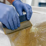 Blue gloved hands using a plastic squeegee to wet out fiberglass cloth with resin inside a white hull, the soaked cloth turning amber and translucent