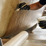 Gloved worker brushing epoxy resin onto fiberglass cloth on a boat hull, digital thermometer reading 23.6 °C and mixing cups on the bench