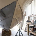 Gloved hand rolling gray epoxy barrier coat onto the underside of a fiberglass hull on jack stands in a metal boat shed, ahead of bottom paint