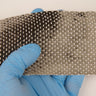 Section of 12K unidirectional carbon fiber tape held up in a blue-gloved hand, light catching the parallel tows and white cross threads