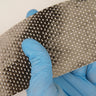 Strip of 11 oz unidirectional carbon fiber tape flexed in a blue-gloved hand, showing parallel 12K tows and finished edges